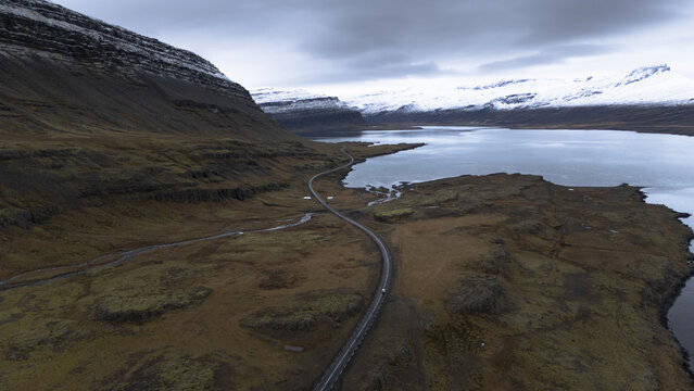 Aerial view of a winding road snakes through the rugged, snow-dusted landscape, contrasting with the serene, reflective waters under a brooding sky, Selfoss, Iceland.