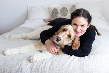 Obraz premium Cute brunette smiling young woman lying down on bed hugging adorable friendly Parti Goldendoodle with white and ginger coat