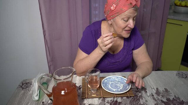 Elderly woman drinks compote from a glass at home. Pour a drink from a carafe. Eats sweet cookies