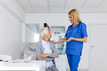 Friendly female doctor in blue scrubs with a stethoscope talking to and comforting an elderly senior patient during a medical consultation in a bright clinic office.