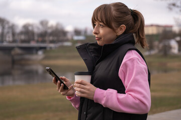 Young woman wearing casual clothes is standing by a river, holding a takeaway coffee cup and using her smartphone, enjoying a peaceful moment outdoors