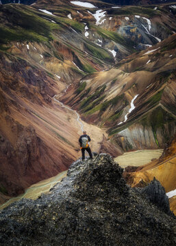 Aerial view of a lone figure standing atop a craggy peak amidst a dramatic landscape of earthy tones and snowy patches, Landmannalaugar, Iceland.