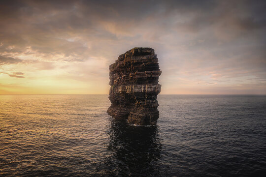 Aerial view of a monolithic sea stack rising from the dark, swirling waters under a sky ablaze with the warm hues of the setting sun, Downpatrick Head, Ireland.