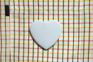 Heart badge mockup. A pristine white heart-shaped badge rests on a multicolored plaid shirt featuring red, yellow, green, and brown stripes, creating a contrast between minimalist design and cheerful 