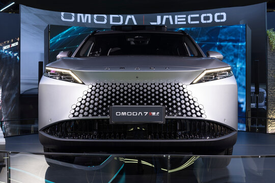 ront view of the Omoda 7 SUV, featuring a three-dimensional geometric grille, slim LED headlights, and metallic finish in an automotive show booth.