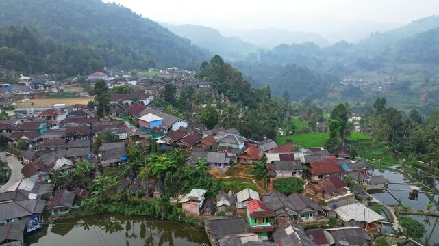 Aerial drone footage of Cisitu village, in Lebak regency, Banten, Java, Indonesia, with many local colorful houses, fish farms and trees, a wide valley with mountains and rice fields, cloudy weather