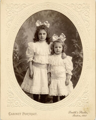 An authentic sepia cabinet card portrait of two sisters from the early 1900s. The girls wear frilly white dresses and large, elegant hair bows.