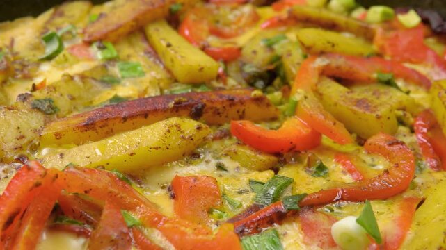 Close-up panning shot across a freshly cooked frittata in a skillet. The camera moves over golden potatoes, fluffy eggs, red peppers, and green onions. An appetizing, ready-to-eat homemade dish