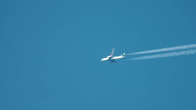 Jet aircraft flying across blue sky at high altitude. Airplane leaving chemical trail forming white contrails, polluting atmosphere. Commercial airliner spreading harmful emissions globally, affecting
