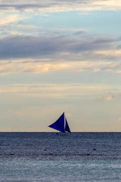  &laquo;The serenity of the open sea: a traditional paraw sailboat with vibrant blue sails glides gracefully across the tranquil waters under a soft, cloud-draped sky &mdash; a picturesque moment of calm and free