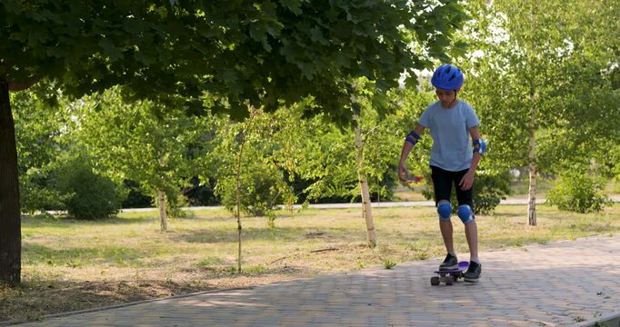 Active boy rides purple skateboard in green park. Little boy wears blue safety helmet and pads. Happy boy enjoys summer sport outdoors. Child skater balances on board on path.