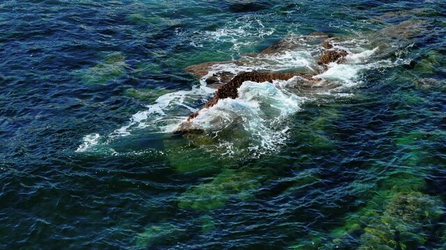 Waves crashing over coral encrusted rocks in clear sea. Surf foaming around jagged reef under turquoise water. Ocean swirling against barnacle covered formation near coast