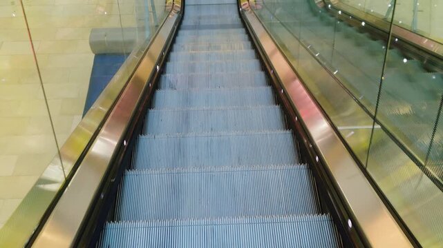 Escalator moving downward inside shopping mall interior with transparent railings. Mechanical steps transporting shoppers between floors in modern retail center. Glass balustrades reflecting lights