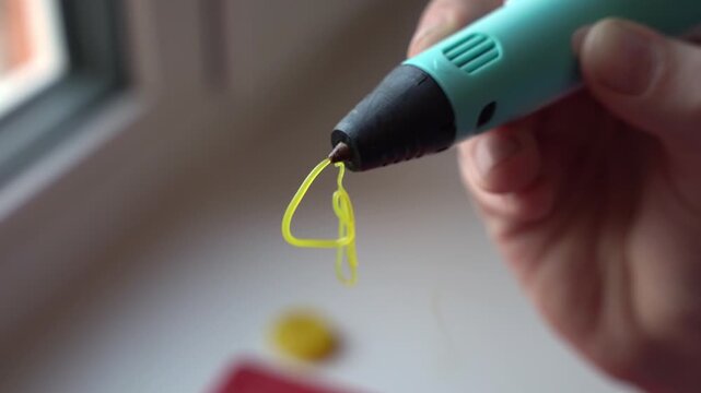 Hand holding a 3D printing pen extruding bright yellow filament in a continuous flow, showcasing the creative process in a well-lit indoor workspace