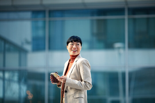 Smiling businesswoman with smartphone and coffee outside modern office