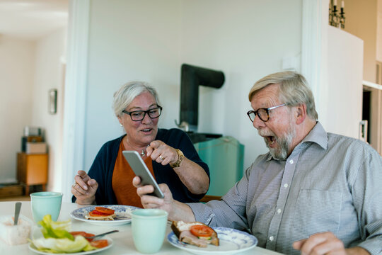 Senior couple reacting to smartphone at home breakfast table