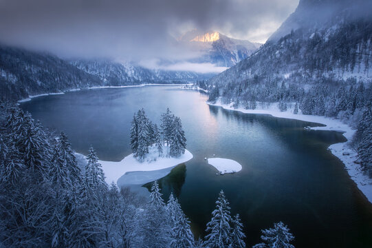 Aerial view of a serene lake cradled by snow-laden forests and majestic mountains under a soft, diffused light, Tarvisio, Friuli-Venezia Giulia, Italy.