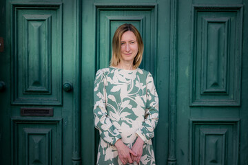 Fototapeta premium A woman stands in front of a large green wooden door, wearing a light patterned dress. She looks calmly at the camera in soft daylight, with a simple urban background.
