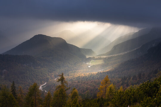 Aerial view of sunbeams pierce through the clouds, illuminating the valley and distant mountains, casting shadows across the landscape, Log pod Mangartom, Slovenia.