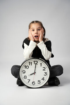 Shocked Young Girl Sitting with Large Wall Clock.  A surprised young girl sitting cross-legged behind a large analog clock showing 8:00, expressing amazement or urgency against a gray background