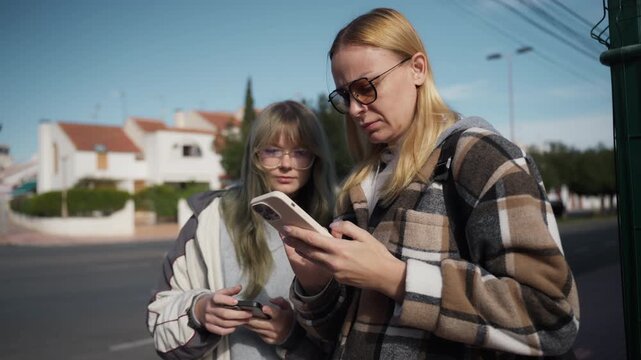 Mother and teenage daughter stand on sunny street, intently use smartphones to search for directions, showcasing their connected yet independent digital lives