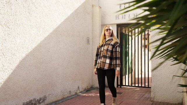 Confident woman wear casual plaid shirt and sunglasses walk out from gated passageway onto sunlit street, enjoy warm weather and urban journey