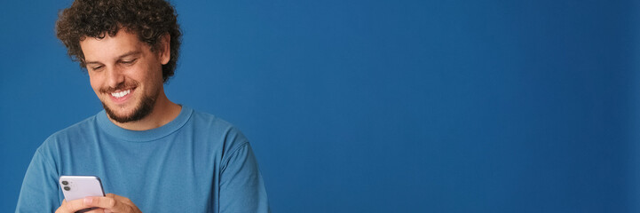 Obraz premium Guy with curly hair dressed in blue t-shirt holds mobile phone in his hands with place for mockup or an ad with copy space on blue background in the studio. Panoramic