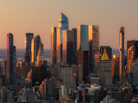 Aerial view of gleaming skyscrapers reflecting the warm hues of the setting sun, casting long shadows over the intricate cityscape, New York, New York, United States.
