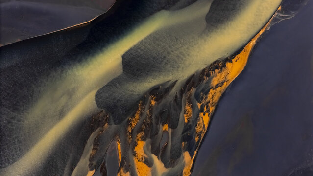 Aerial view of braided river channels intertwine like veins of gold and silver, carving through the stark, volcanic landscape, creating a mesmerizing tapestry of light and shadow, Selfoss, Iceland.