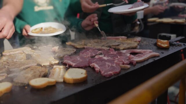 People enjoy barbecue party, serving sizzling pork steak, bacon, and bread from  large outdoor plancha grill, surrounded by steam and inviting aroma of cooking food