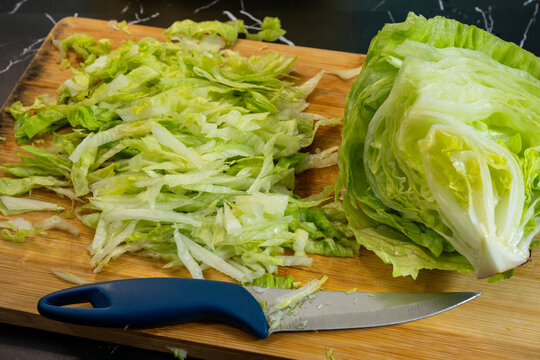 a wooden cutting board featuring a pile of shredded green lettuce next to a partially sliced head of iceberg lettuce and a knife with a blue handle.