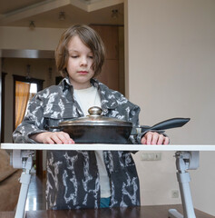 Young boy standing with frying pan on table. Kid waiting for dinner in kitchen