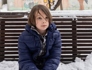 Portrait of boy sitting on snowy bench. Thoughtful child in winter park. Cold weather clothing