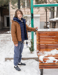 Young boy standing by snowy bench in winter. Kid in warm brown jacket outdoors