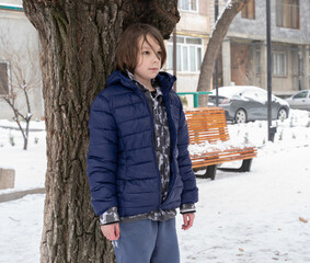 Boy standing near tree in winter park. Child in blue jacket outdoors. Snowy city street scene