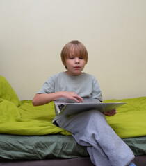 Young boy sitting on bed using laptop. Child learning or playing online at home