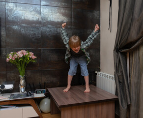 Playful boy standing on table with arms raised in victory. Naughty child having fun in living room