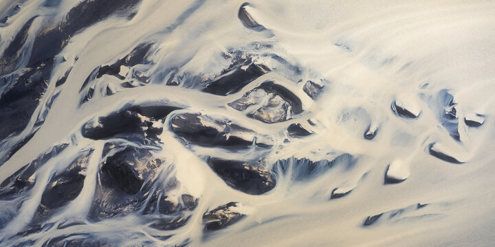 Aerial view of glacial river patterns create intricate designs across the landscape, as icy water flows around dark rocks, Iceland.