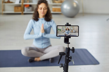 Fototapeta premium Woman yoga influencer and online content creator films calming zen meditation lesson tutorial class video in phone app, sharing wellbeing and self care guidance. Device screen and tripod in soft focus