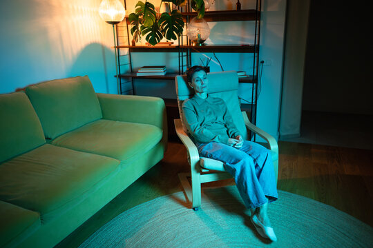 Loneliness and depression of elder people, senior woman sat in chair at home in rgb light looking with apathy and sadness, life crossroads drama and mental exhaustion caused by drama in relationships.