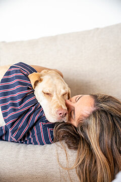 A woman is laying on a couch with her dog on her lap. The dog is wearing a striped shirt and he is sleeping. The woman is looking at the camera and she is smiling