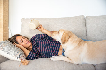 A woman is laying on a couch with a dog on her lap. The dog is playing with a cup, and the woman is...