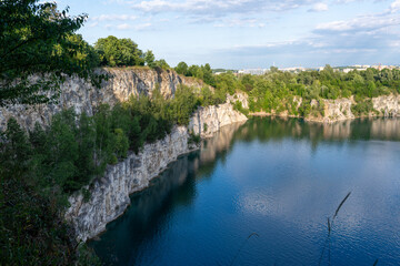 Fototapeta premium Wide landscape of zakrzowek quarry lake in Krakow, Poland, showing limestone cliffs, green trees, and calm blue water under a bright sky, representing urban nature escape, summer recreation, hiking vi