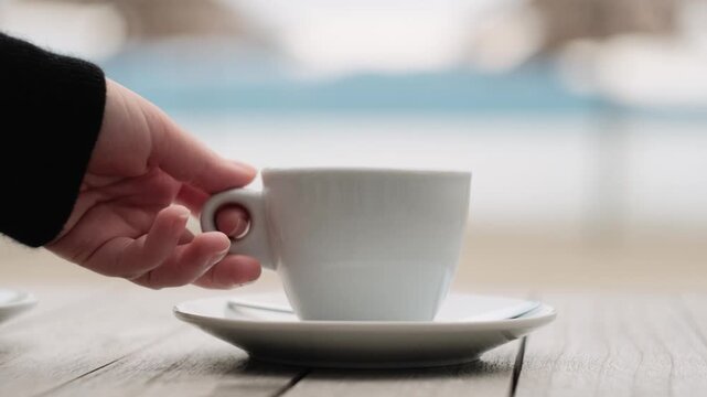 Female hand placing a cup of coffee on a saucer on a wooden table. Close-up lifestyle scene with soft natural light, cozy coffee break, slow living and daily routine concept.