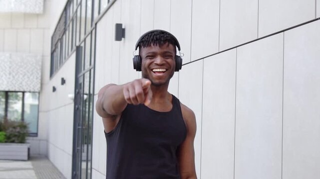 African American man with headphones mocks and laughs while pointing at the camera, sporty guy accuses and ridicules you on city street