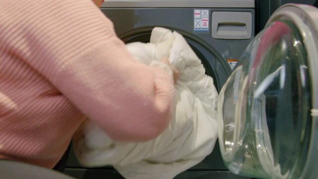 Individual in pink sweater loads a large white comforter into a front-loading washing machine at a self-service laundromat with modern appliances and bright lighting