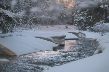 Cold winter river in a snowy forest with flowing water and icy edges. Natural winter scene showing contrast between frozen snow and moving stream. Concept of cold climate, seasonal change