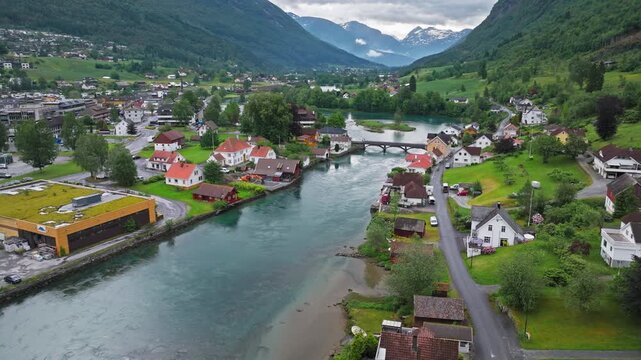 Drone footage moving above Stryn town center along a calm river channel. Serene summer mood captures houses, roads, and mountain backdrop.