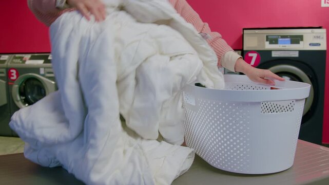 Individual prepares a large white comforter for washing at a self-service laundromat, placing it into a white laundry basket on a folding table