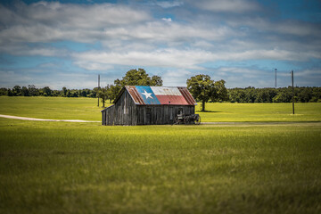 Old Rustic Barn With Texas Flag Painted on Roof With Old Wagon Nearby In Green Field in Rural Town in Texas © Lone Star Stock
